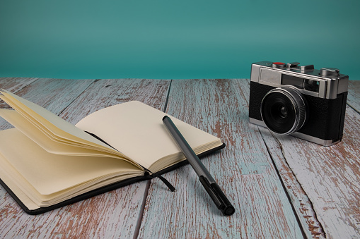 Notebook with a pen and a photo camera, on a wooden table with a light blue background. Free time concept.