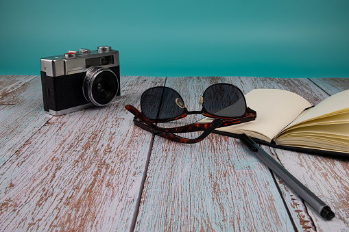 Camera, sunglasses with a notebook and a pen on top of a wooden table, on green background. Vacation concept.