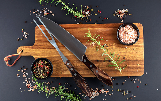 Wooden cutting board, rosemary and spices with fork and knife carving set on a dark background
