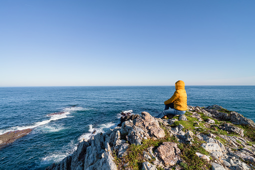 Coastal landscape of rocks and sea with woman sitting looking at the sea with yellow hooded jacket. Copy space.