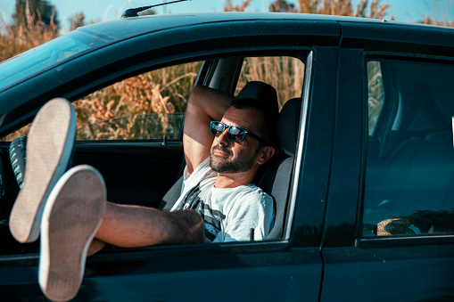 A photo of attractive man wearing sunglasses. Happy young male is enjoying road trip in car. He is wearing casuals.