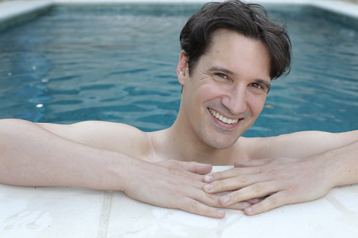 A shirtless handsome man in his 30s is smiling in a swimming pool. He is inside the water and looks cheerful.