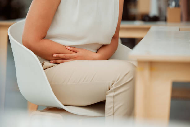 primer plano de una mujer de negocios de raza mixta que sostiene dolor de barriga mientras se siente enferma con calambres estomacales menstruales y dolor de vientre en una oficina. empleado hambriento en período enfermándose, hinchado e incómodo con do - holding belly fotografías e imágenes de stock