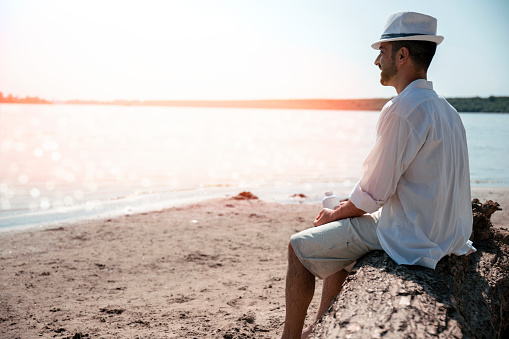Bearded mature Man in white shirt and hat, sitting on the tree trunk at the sand beach, enjoying in summer sunset. Copy space