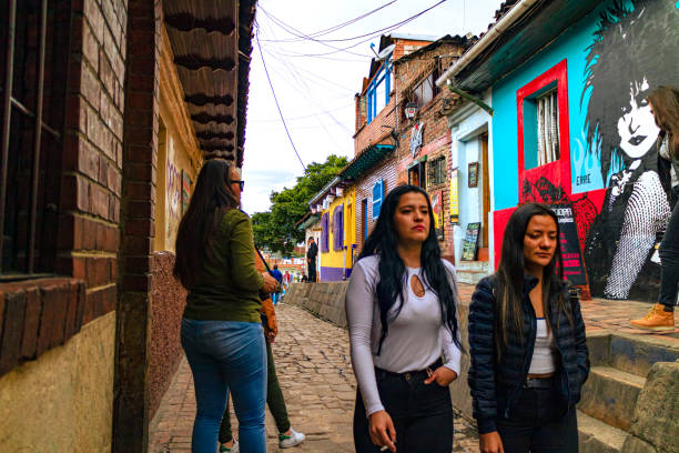 Bogota, Colombia - Local Colombians On The Calle del Embudo, In The Historic La Candelaria District Of The Andes Capital City In South America. Bogota, Colombia - October 20, 2019: Local Colombian women walk up the narrow Calle del Embudo, one of the most colorful streets in the historic La Candelaria district of Bogotá, the Andean capital city of the South American country of Colombia. The street leads to the Chorro de Quevedo, the plaza where it is believed the Spanish Conquistador, Gonzalo Jiménez de Quesada founded the city in 1538. Many street facing walls in this area are painted with either street art or the legends of the pre-Colombian era, in the vibrant colours of Colombia. The altitude at street level is 8,660 feet above mean sea level. Photo shot on an overcast morning; horizontal format. calle del embudo stock pictures, royalty-free photos & images