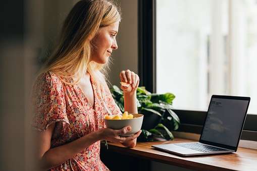 Blonde woman eating fruit salad while browsing her social media on her laptop.