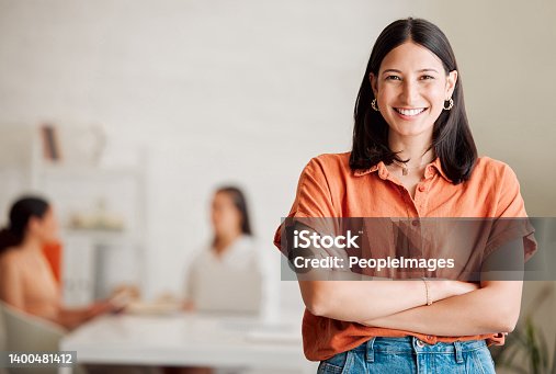 istock Portrait of one confident young hispanic business woman standing with arms crossed in an office with her colleagues in the background. Ambitious entrepreneur and determined leader ready for success in a creative startup agency 1400481412