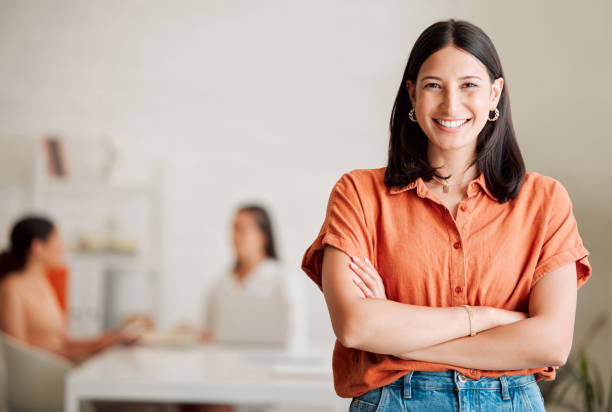 Portrait of one confident young hispanic business woman standing with arms crossed in an office with her colleagues in the background. Ambitious entrepreneur and determined leader ready for success in a creative startup agency stock photo