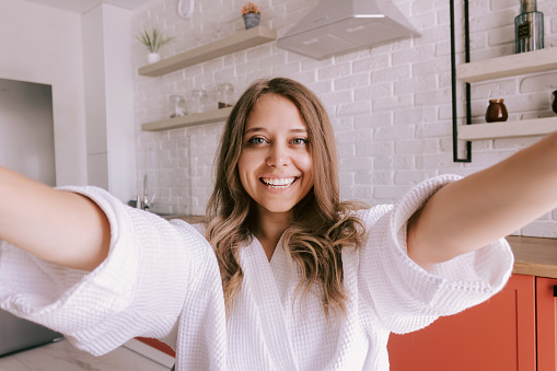 A young beautiful caucasian smiling blonde woman in a white robe takes a selfie reaching out to hug in the morning on background of the kitchen