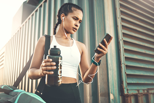 One fit young hispanic woman listening to music with earphones on cellphone and carrying sports tog bag and water bottle for exercise outdoors. Female athlete texting and choosing songs for playlist while walking to or from gym