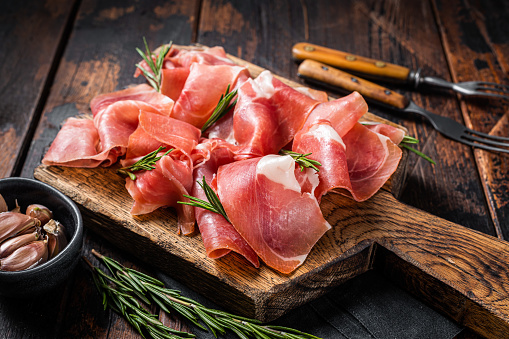Slices of jamon serrano ham or prosciutto crudo parma on wooden board with rosemary. Wooden background. Top view.