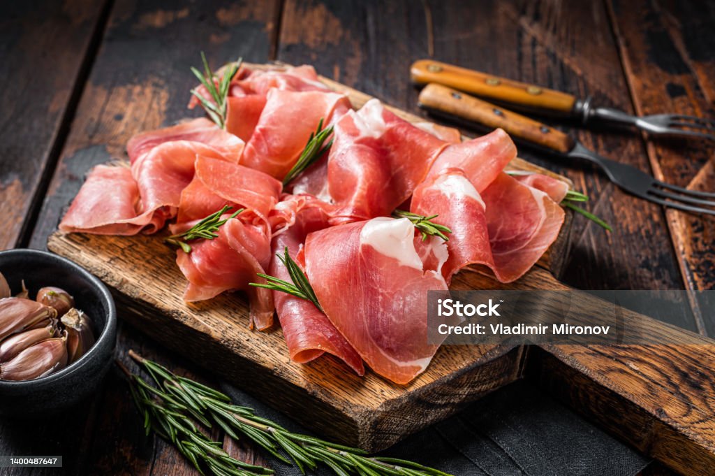 Slices of jamon serrano ham or prosciutto crudo parma on wooden board with rosemary. Wooden background. Top view Slices of jamon serrano ham or prosciutto crudo parma on wooden board with rosemary. Wooden background. Top view. Prosciutto Stock Photo