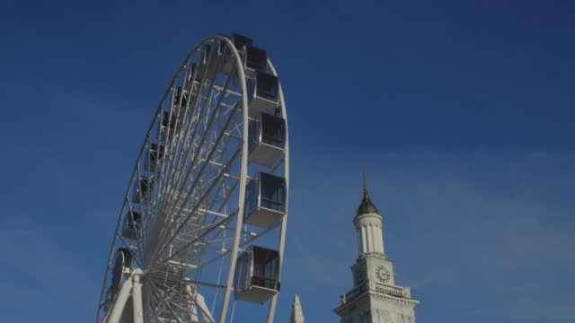 Details of a beautiful city square. A spinning Ferris wheel and a bell tower against a blue cloudless sky.