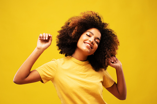 lifestyle, emotion and people concept: young african woman portrait with big afro curly hair on yellow background dancing and with hairstyle flying in air