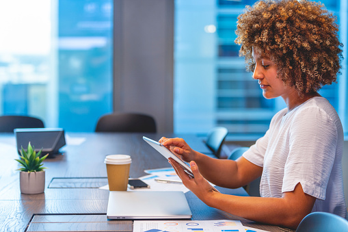 A young female businesswoman looking at her digital tablet . She is sitting at a table in a modern office. She looks relaxed and could be surfing the internet or shopping online. She looks like the business owner or manager. There is a laptop and paperwork on the table.