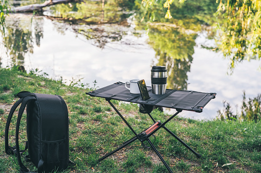 Camping table with cup of coffee and smartphone outdoors. Everyday technology. Picturesque landscape. Recreation and travel concept