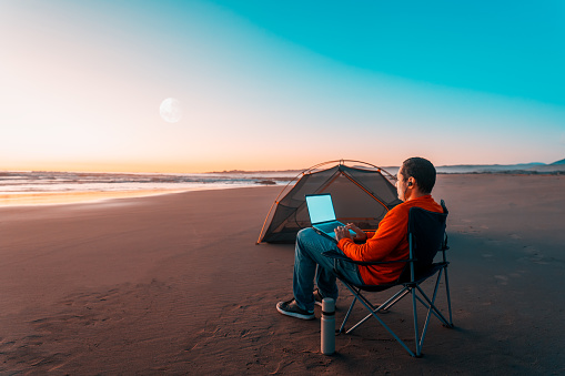 digital nomad sitting alone on the shore of the beach with a laptop working next to the tent at sunset
