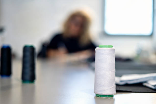 Closeup of spools of thread on work table, colorful threads. Out of focus background of woman working on her sewing designs in her small business.
