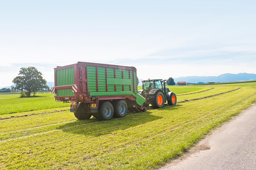 Tractor with a loading wagon harvesting hay on a huge meadow in the Bavarian Alps, Germany.