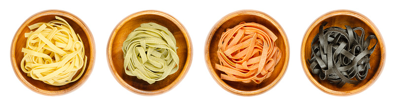 Tagliatelle pasta, in four different colors, twisted into nests, in wooden bowls. Uncooked and dried traditional Italian type of egg pasta. Long flat ribbons, classic, spinach, tomatoes and squid ink.