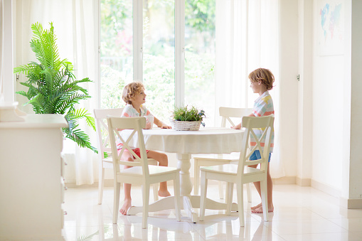 Kids eating breakfast in white kitchen. Children eat sandwich and drink juice on sunny morning. Family at home. Boy cooking lunch. House dining room interior. Child healthy nutrition.