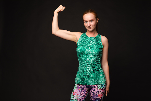Studio portrait of sporty woman in bright colorful sportswear showing strong biceps on black background. Sport concept.