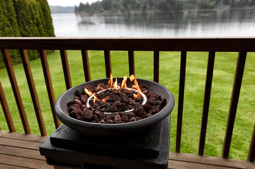 Burning gas fire pit, natural lava stones, on outdoor home deck with lake and woods in the background
