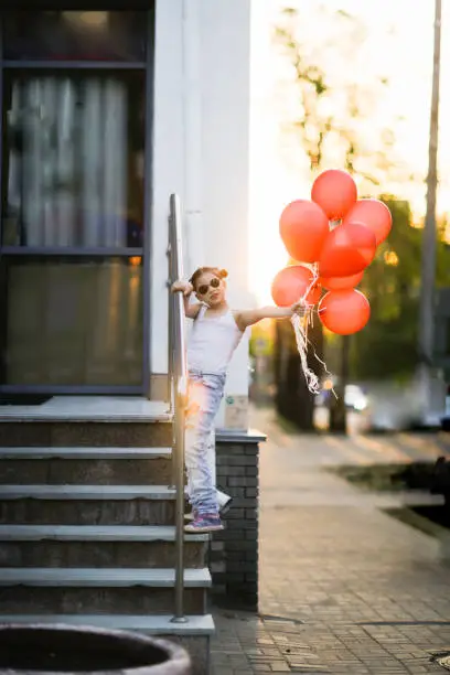 Photo of European child preteen girl in torn jeans with red balloons on stairs sunny summer day. Child in sunglasses at sunset on city streets, generation Z, happy birthday