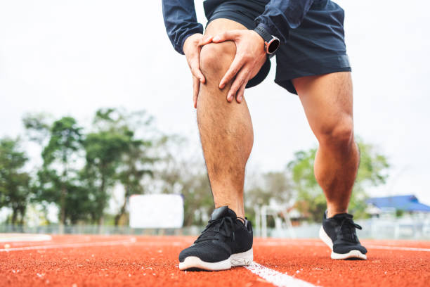 o jovem corredor está com o joelho dolorido porque correu muito tempo. homem se exercitando até a lesão. supertreinado. jovem acidente de esporte correndo na pista - cramp - fotografias e filmes do acervo