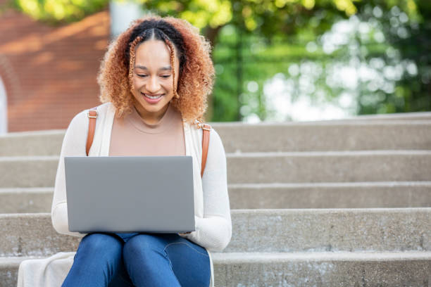 istock Beautiful young woman uses laptop computer while sitting on steps on university campus 1400534329