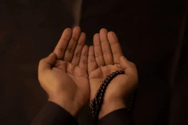 Muslim men raise their hands to pray with a Tasbeeh on Dark background, indoors. Focus on hands.