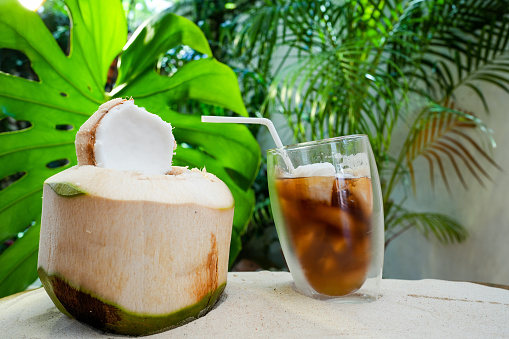 Iced Coconut Coffee In Double Wall Glass On sand Against Green Leaf Background.