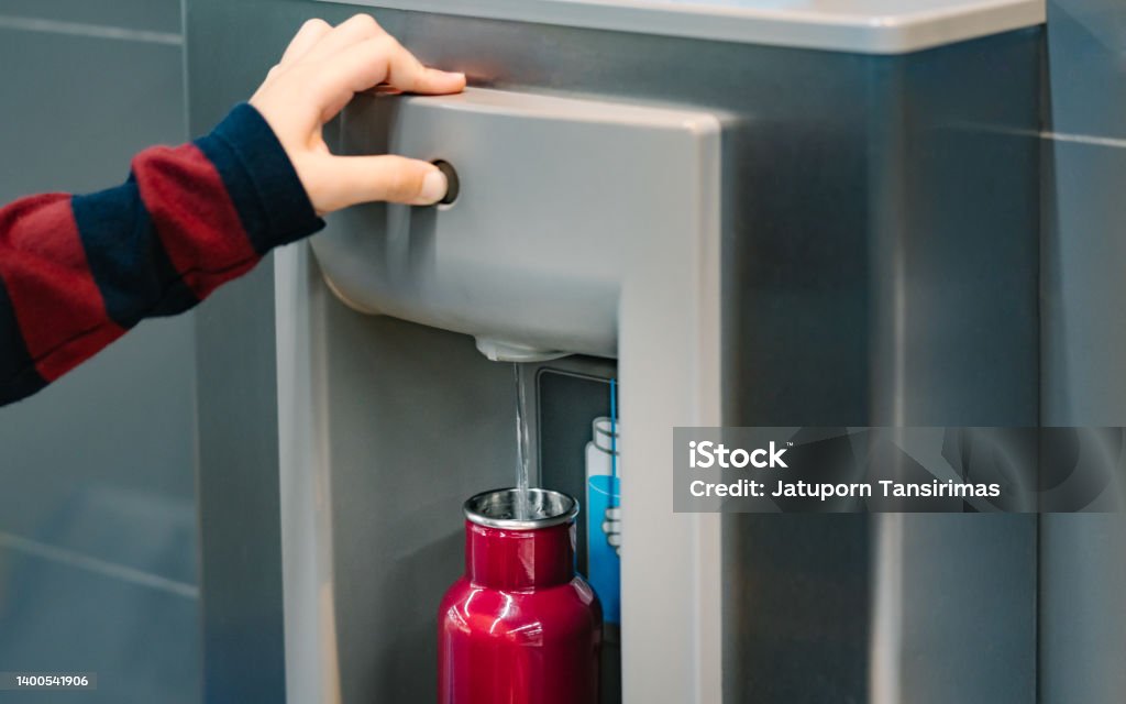 Close up of hand pressing button of drinking water filling station at the airport Close up of hand with long sleeve red blue stripe shirt pressing button of drinking water filling station at the Airport to refill red insulated reusable water bottle. Water Bottle Stock Photo