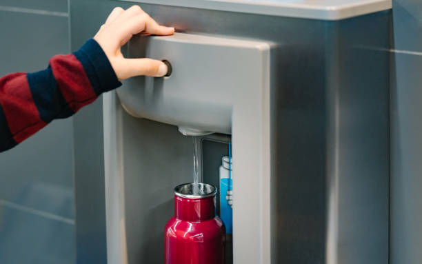 Close up of hand pressing button of drinking water filling station at the airport Close up of hand with long sleeve red blue stripe shirt pressing button of drinking water filling station at the Airport to refill red insulated reusable water bottle. filling stock pictures, royalty-free photos & images