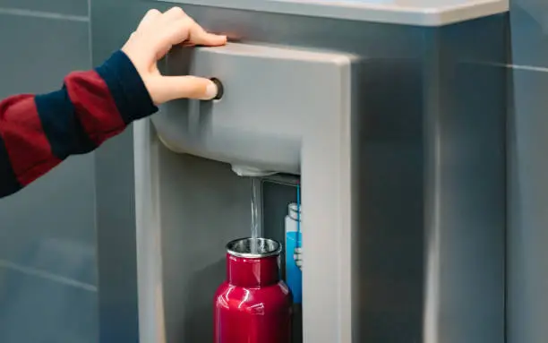 Close up of hand with long sleeve red blue stripe shirt pressing button of drinking water filling station at the Airport to refill red insulated reusable water bottle.