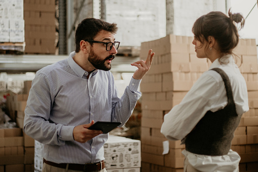 Image of an industrial manager having a conflict with a female worker, explaining what the problem is, while both standing in a big warehouse. He's a bearded guy with eyeglasses on and she's a brunet with bangs, both formally dressed