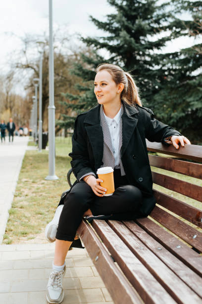 istock Charming young woman in stylish coat resting on bench during coffee break outdoors. Smiling elegant lady holding disposable coffee cup and looking away in park 1400544889