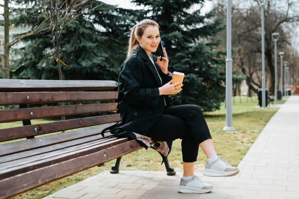 istock Stylish positive young woman calling on mobile phone during coffee break while sitting on park bench. Smiling pretty woman in coat talking on smartphone and holding cup of coffee looking away outdoors 1400544894