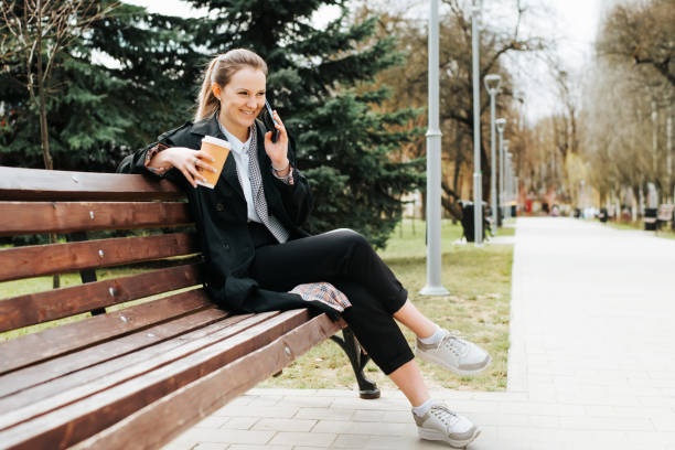 istock Positive fashionable young woman laughing while talking on smartphone while sitting on park bench. Smiling pretty woman wearing stylish coat calling on mobile phone during outdoor coffee break 1400544941