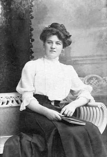 Vintage monochrome photo scan, ca. 1912. Young woman with victorian hairdo and fashionable high-necked blouse sitting on a classic bench.