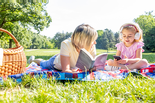 Joyful child having smart phone and headphones while sitting by a parent reading a book on a picnic