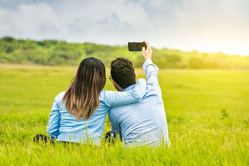 Young couple in love taking a selfie in the field, People in love taking selfies in the field with their smartphone, Smiling couple in love sitting on the grass taking selfies