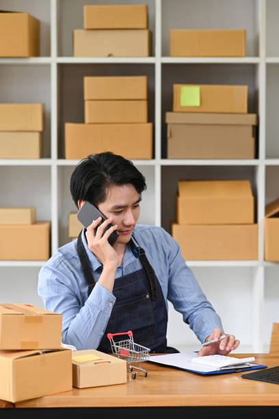 istock Young male small business owner  having cell phone conversation for confirming orders from customer. Online selling, e-commerce concept. 1400556689