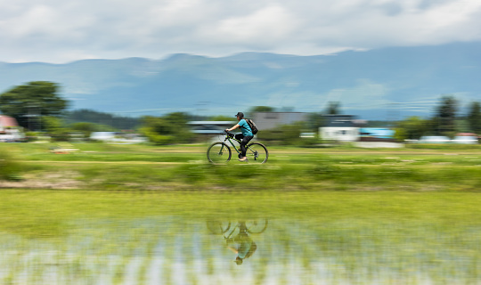 A woman riding by rice fields in the countryside with her reflection on the foreground water and mountains behind. The entire scene shows speed blur except for the woman on the bike.