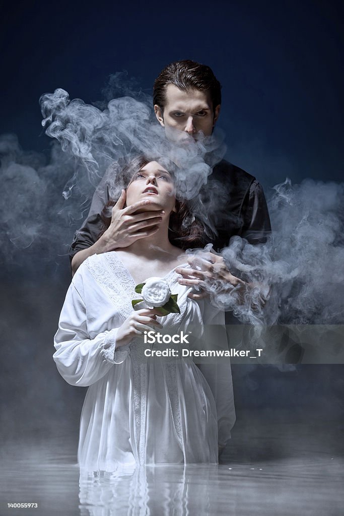 Hades & Persephone: The Seduction Dark-haired, sinister, masterful man dressed all in black standing behind youthful virginal girl in pure white dress holding a white flower looking upwards. He holds her by the throat gazing at camera mysteriously and seductively. Both standing in pale waters with white mist against a midnight background. Fantasy Stock Photo