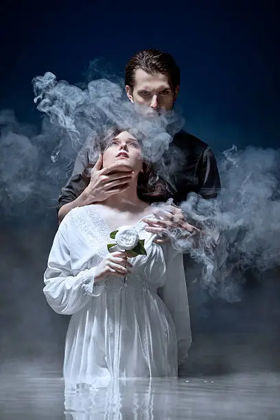 Dark-haired, sinister, masterful man dressed all in black standing behind youthful virginal girl in pure white dress holding a white flower looking upwards. He holds her by the throat gazing at camera mysteriously and seductively. Both standing in pale waters with white mist against a midnight background.