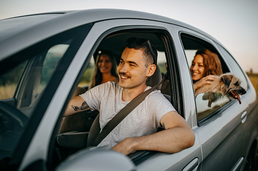 Group of friends going on road trip. Thy are sitting in the car, dog is looking outside the window. They are driving and enjoy talking and laughing