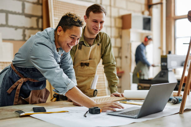 Carpenters Designing in Shop Warm toned portrait of animated carpenters using laptop and discussing furniture designs in workshop small business stock pictures, royalty-free photos & images