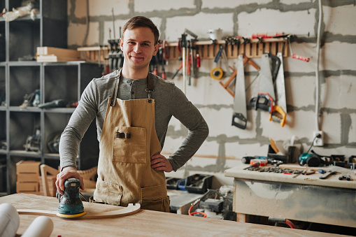 Warm toned shot of young carpenter smiling at camera while building furniture in workshop, copy space