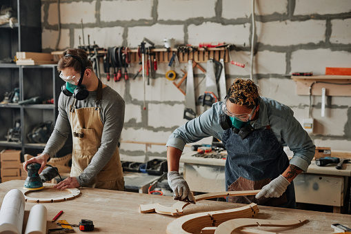 Warm toned portrait of two carpenters in protective gear working with wood while building handicraft furniture in workshop